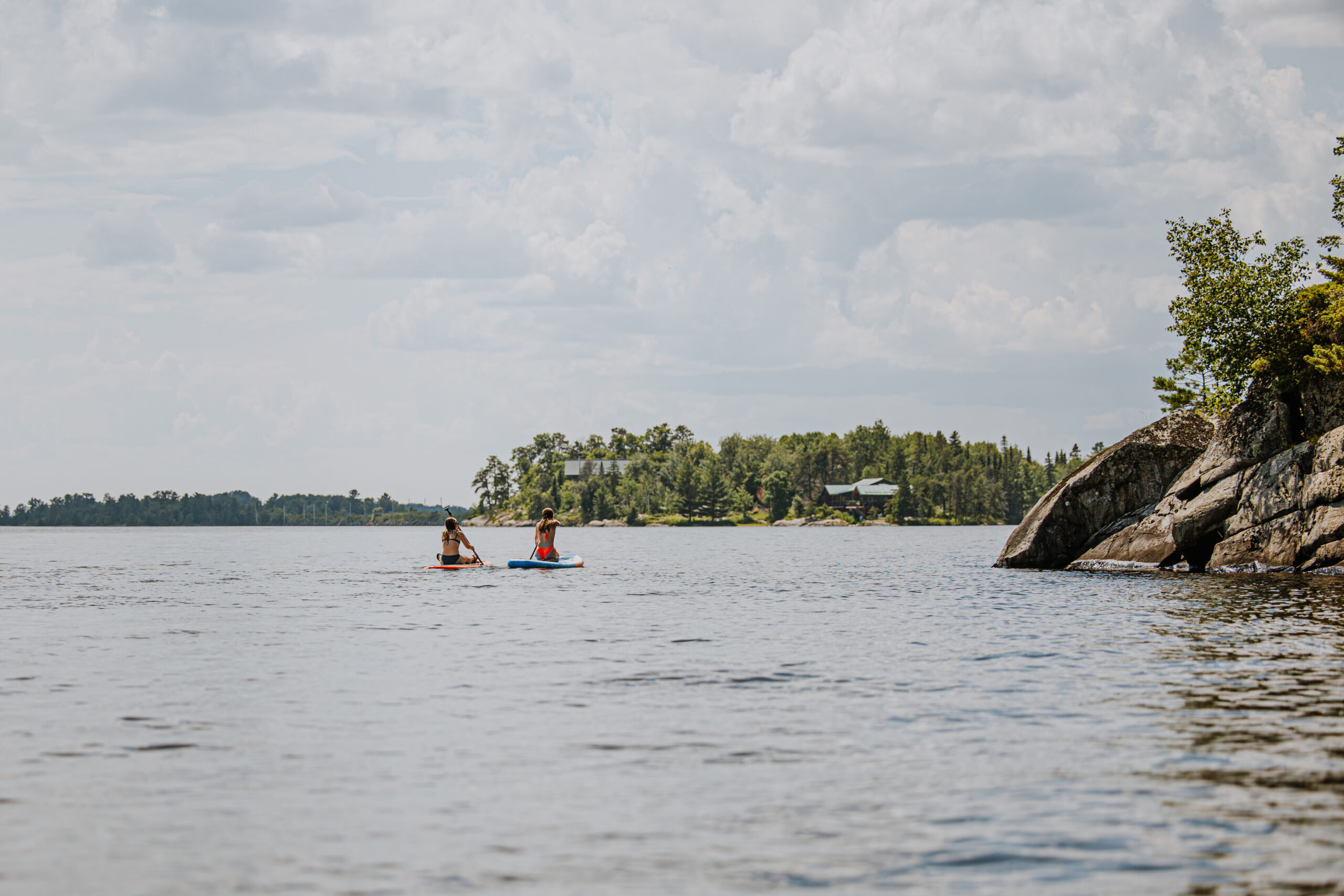 Two girls off on the distance on paddlebaords in Rainy lake. Large granite boulders off to the right. Sunny day with big puffy clouds in the sky.